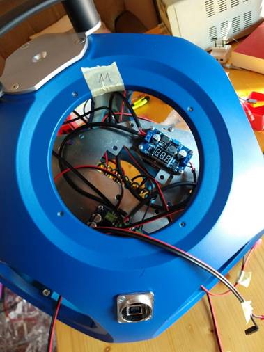 A piece of the blue plastic shell forming the chassis of the dodecahedron. The speaker driver itself is missing, and through its mounting hole the internal electronics can be seen, in particular a blue board controlling relays, with a 3-digit 7-segment display, and the green relay board.